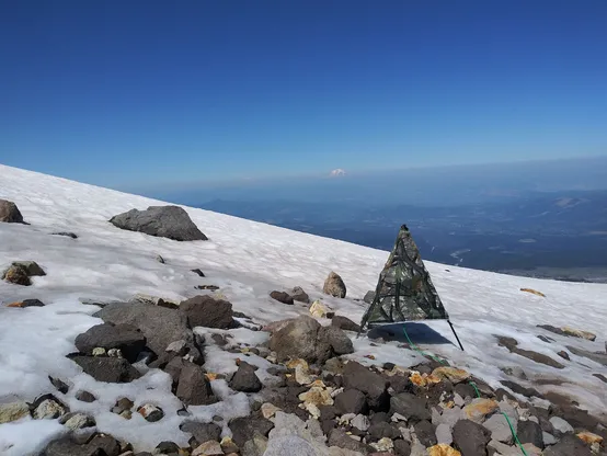 A tripod holding a microphone standing on the icy slope of Mt. Hood. It is a clear day with blue sky and in the far distance is Mt. Rainier slightly obscured by wildfire smoke.