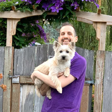 The author smiling while holding a small terrier. The color of his shirt coordinates with flowers in the background.