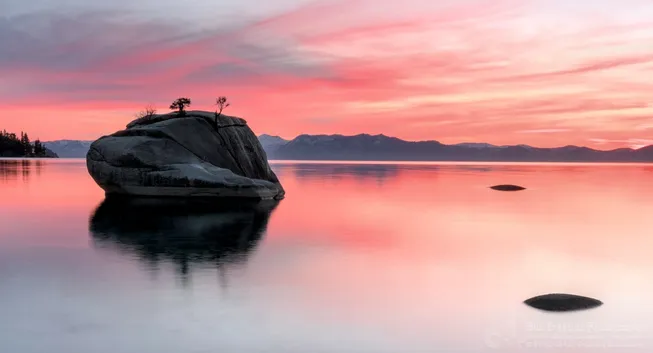 Bonsai Rock in Lake Tahoe, Nevada, is a large granite boulder just off the eastern shoreline. It's name comes from the two small "bonsai" trees growing in cracks at the top of the rock. The photo shows the rock in a mirror still lake with a red-orange sunset and distant mountain peaks in the background. (12/29/2017).