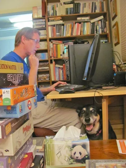 Man working at a computer on a desk, surrounded by books and board games, with a nervous dog peeking out from under the desk.