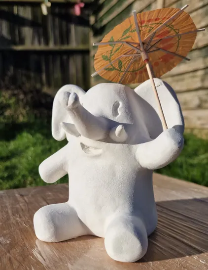 Clay model of the Mastodon mascot, sitting down on a wooden table in the sun and holding a cocktail umbrella.