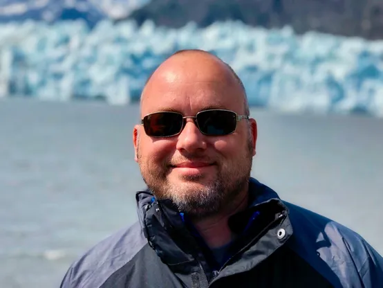 Profile photo of R Scott Jones, on a boat in Glacier Bay National Park in Alaska as he finished his lifelong quest to visit all 419 National Park units in the US. He’s smiling in a jacket with sunglasses on and the edge of a glacier in the background.