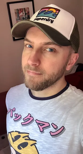 Selfie of a bearded guy in a superdry cap. He wears a t-shirt with a picture of PacMan on it, and the Japanese katakana for "pakkuman."