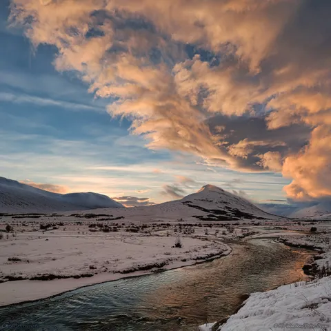 A snow scene, with mountains in the distance. A river runs from centre bottom of the shot, winding its way towards the right then to the left. The banks are covered in snow and ice. Above a distant pointed mountain, a bank of cloud angles up from the right centre to the top left in a triangular wedge. It's lit by the rising sun in soft shades of orange and pink. The sky above it is hazy with icy cloud, and the flowing river below reflects the sunrise. The overall mood is peaceful and awe-inspiring.