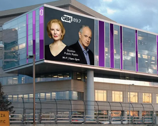 A modern building with a digital billboard with the faces of Boston Public Radio hosts Jim Braude and Margery Eagan, reading Boston Public Radio, 11AM-2PM