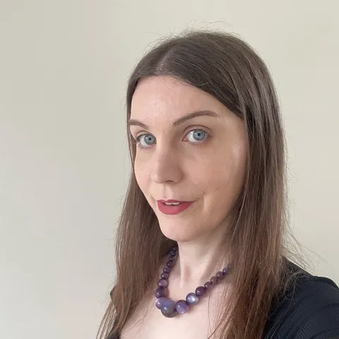 A photo of me, a 41-year-old white woman with blue eyes and long brown hair parted in the centre, wearing a black top and a necklace of large purple beads