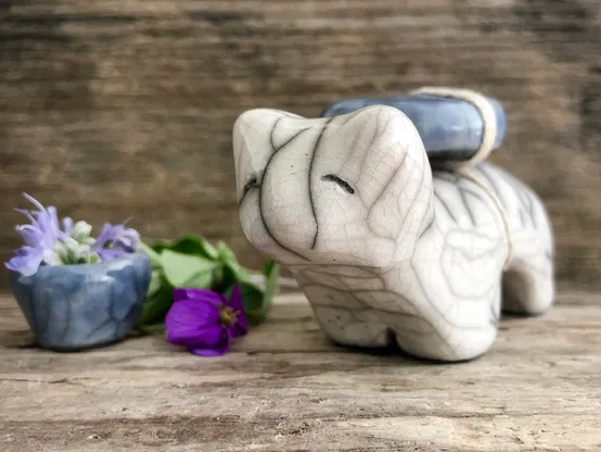 on a background of grey-brown driftwood, a raku ceramic white bear sculpture. it has a very sweet face, and there is a blue crescent moon tied to its back. it has a little blue cup filled with rosemary flowers next to it on the left, along with a violet and a sprig of oregano.