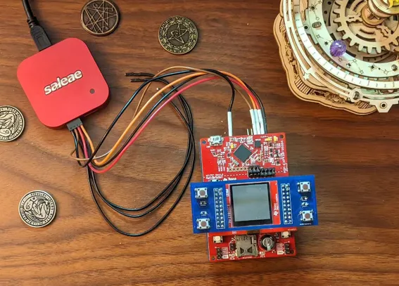 Photograph of a red-and-blue electronic gizmo on a wooden desktop surface. There are some prop coins featuring lovecraftian images scattered around. A laser-cut wooden orrery is visible in one corner. A bundle of wires connects the gizmo to a red "saleae" logic analyzer, which has a USB cable running out of frame attached to it.