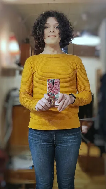 Mirror selfie taken from below the chest, wearing a yellow shirt and blue jeans, head held high, smiling and looking down into the camera