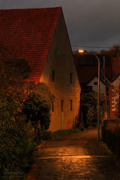 Photo in warm colors of a large, old, red tile roofed stone barn, illuminated by a street lamp.