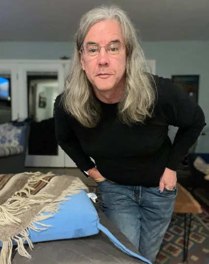 Photo of myself. I have my hands partially in my pockets (blue nail polish), wearing a black long sleeve t-shirt and blue jeans. My hair is down 6-7 inches below my chin. I'm wearing glasses, and I'm leaning over and peering into the camera with a half smile on my face.