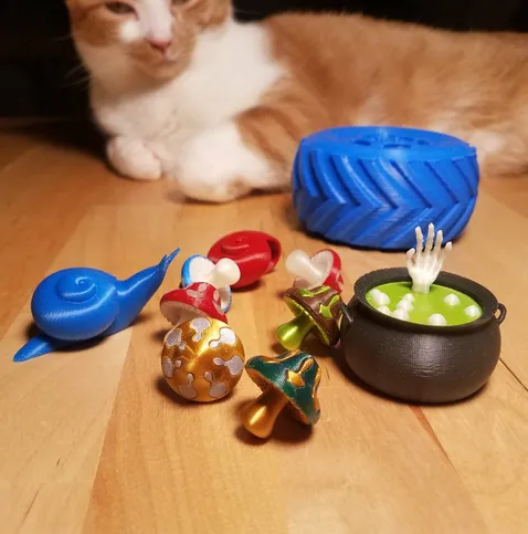 A variety of 3D printed plastic models that I made:  a bubbling cauldron with a  skeleton hand reaching out, various colorful mushrooms, a snail, and a monster truck wheel.  A normal kitten is in the background for scale - the objects are relatively small.