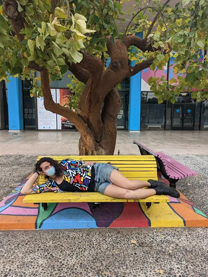 A picture of me in an artsy patterned button down lying on a yellow bench beneath a tree. There is a cement border around the tree with an artsy pattern similar to the one on my shirt.