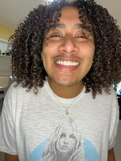 Non-binary person with shoulder length curly hair wearing a white Britney Spears T-shirt smiling into the camera. 