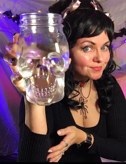 Goretta Synn, a dark haired woman sporting a large spider necklace holds up a skull shaped mason jar full of water to encourage hydration.