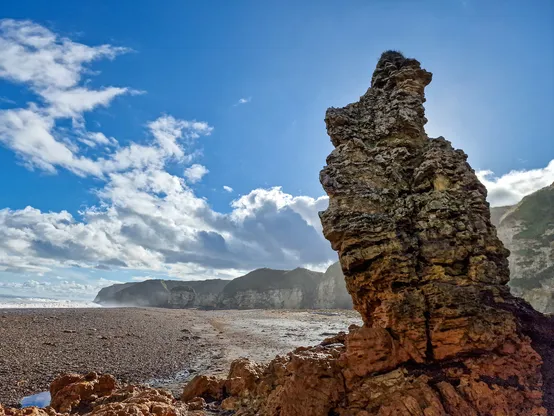 Image of a rock formation on a rocky beach with the sea and cliffs in the background. The sky is blue with some white fluffy clouds.