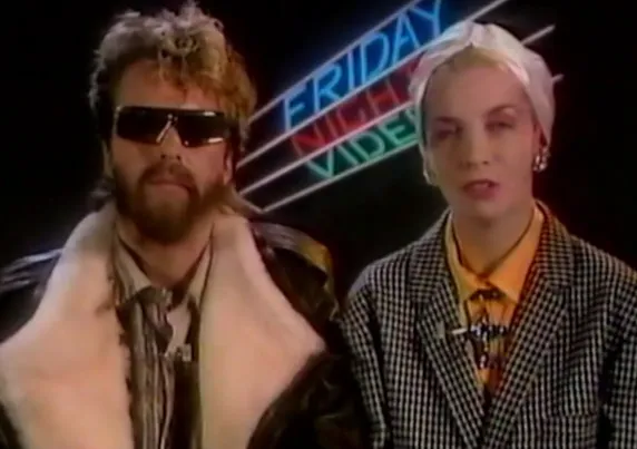 Two people in 1980s fashion with a "Friday Night Videos" neon sign in the background.