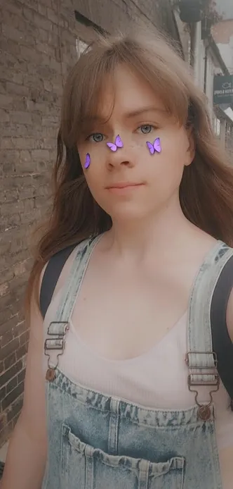 selfie of a girl with dark blonde hair, a pink bralette and dungarees on. there's a Snapchat filter adding butterflies and freckles