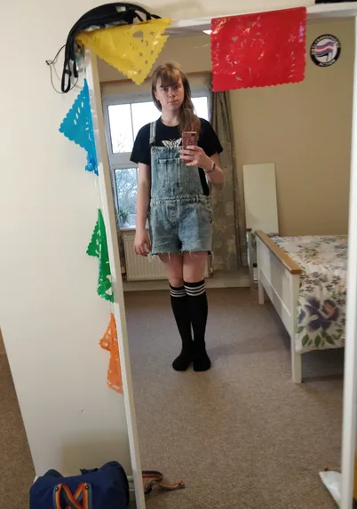 full length mirror selfie of a girl in black thigh highs and top, with blue denim dungarees over them. in frame is an "anti transphobic action" sticker and a bag with rainbow straps. 