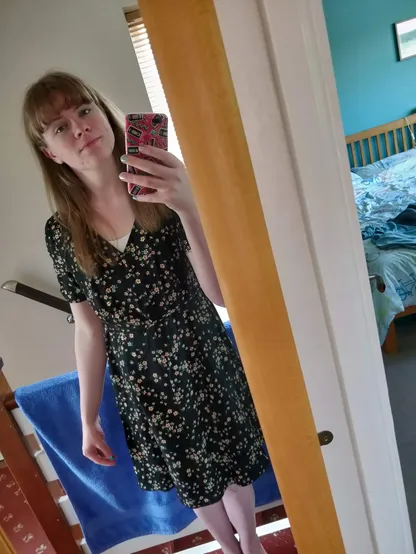 mirror selfie of a girl with dark blonde hair in a black floral dress. her nails are painted poorly in the colours of the trans flag