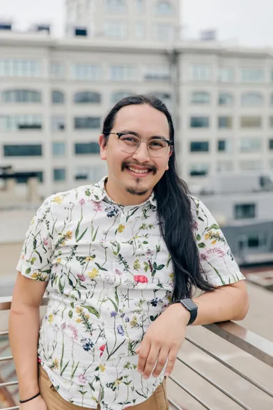 Portrait of me leaning on a railing on a rooftop. I'm a mixed Asian-Caucasian man with wire rimmed round glasses and long black hair in my thirties. I'm wearing a white shirt with botanical flower print.