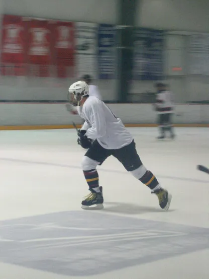 A man playing ice hockey, mid-stride with stick held off the ice at waist level. Wearing a white jersey, helmet, and socks, with black skates, gloves, and pants.