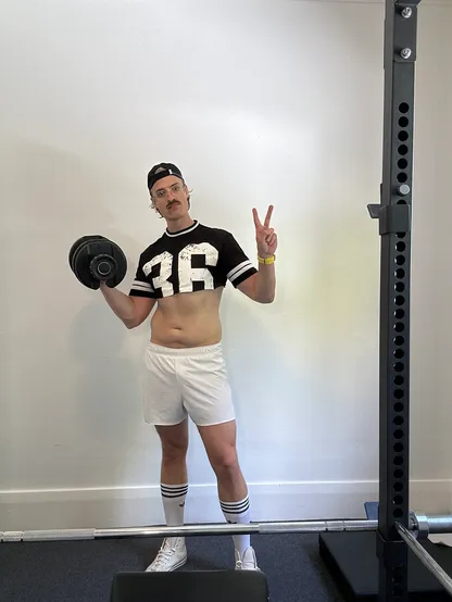 A man in while shorts, black jersey crop top, and a backwards cap holding a dumbbell and giving the peace sign