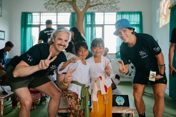 Two men and two young girls smile and give the peace sign during a school visit for the Bali Children’s Foundation. The two young girls are dressed in traditional Balinese clothes 