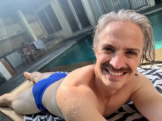 Selfie of a man lying on his stomach by a pool in a blue speedo