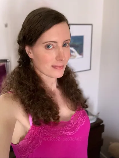 A quarter-angle profile photo of Eva Winterschön, standing in a former bedroom circa 2022. She's wearing a pink cami-top and her long curly hair is set in pig-tails, blue eyes looking into the camera, with a slightly smirking smile.