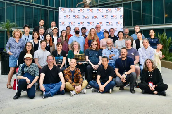 Group photo of 36 people of mixed gender, age and race posing in front of a white backdrop with repeated W30C logos amid palm trees in a courtyard