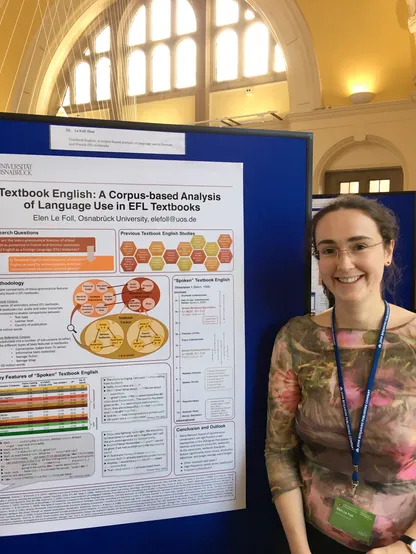 Photo of a white woman with glasses in her 30s smiling, wearing a floral top and a conference badge. She is standing in front of an academic poster entitled: Textbook English: A Corpus-Based Analysis of Language Use in EFL Textbooks.