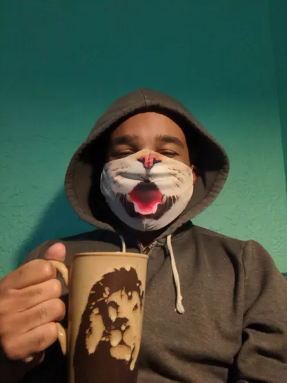 Black man wearing a hoodie and a cat mask with an open mouth holding a mug of coffee