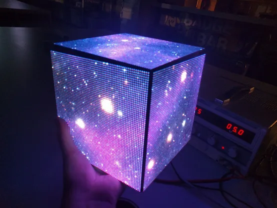 In my hand, I am holding a cube about the size of a pumpkin. It consists of LED pixelated panels displaying a still image of a purple deep space star field. The surroundings are dark. The light is shining on my hand and arm.