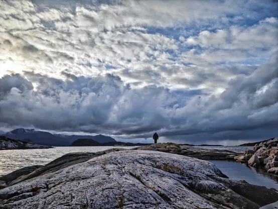 A silhouette standing in front of a cloudy Norwegian fjord, looking towards the sea. A picture of natural power and silent unity with the elements.