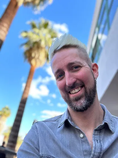 Selfie of a white man with bleach blond hair and a graying dark beard, wearing a button up shirt and looking slightly downward at the camera. There are Palm Trees and partly cloudy skies in the background.