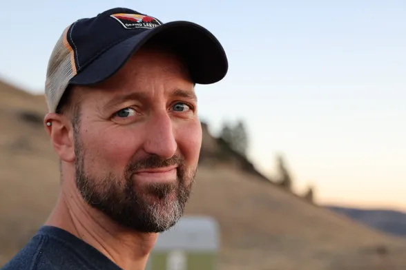 Portrait of a white man with a graying beard and wearing a baseball cap and a slight smile on his face. The blurred background indicates a rural hilltop with some trees and a small cabin in the distance.