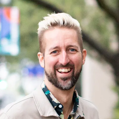 Semi-professional portrait from chest up of a white man with bleach blond hair and graying beard, wearing a floral button up shirt and tan jacket. He is smiling widely.