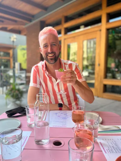 A white man with a dark beard sports cotton candy pink hair. He is sitting across a pink table from the photographer on a restaurant patio, is holding up a pink cocktail, and is wearing a pink and white vertical striped shirt, along with a big smile on his face. The table has menus with pink accents and two glasses of rosé wine.