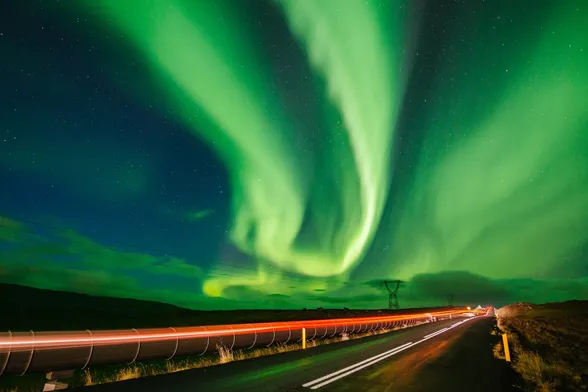 Bright green aurora over a road in Iceland. A car has gone through the photo leaving a trail of red light.