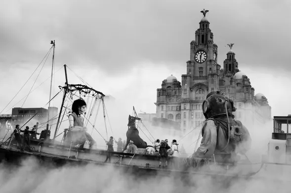 On a boat passing the Liver Building are giant marionettes. A young girl, a dog and their Uncle who is wearing a vintage diving helmet.