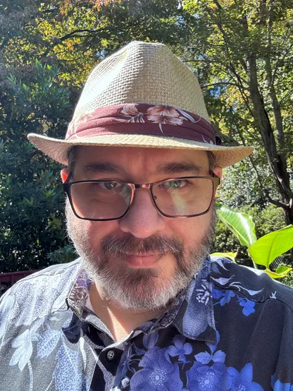 Selfie of Magnus KI4OTK. He is a 50-something white male-presenting person. He has green eyes, salt & pepper beard, glasses. He is wearing a straw fedora and floral print shirt. Background is lush trees and potted outdoor plants. 