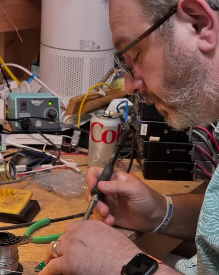 Magnus KI4OTK soldering on a small PC board concealed from view by his left hand. There's a messy work bench in the shack with a stack of old taxi radios in the background, an open can of Diet Coke, and a Weller soldering station.