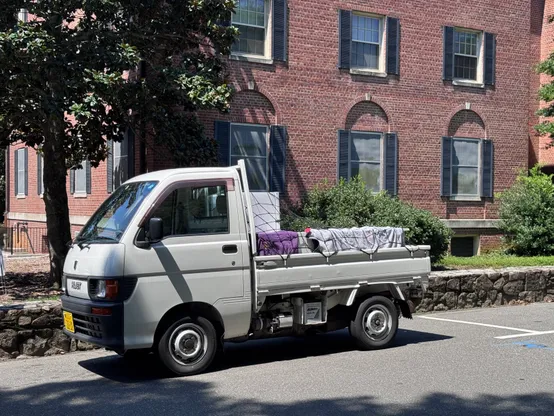 Outdoor photo of a 1998 Daihatsu Hit S110P. This is a Japanese Domestic Market "kei" truck which is very tiny but also very practical. It is parked in front of a university dormitory building, and the load bed is packed with student belongings to move into the dorm. It's the cutest truck known in the QTH.