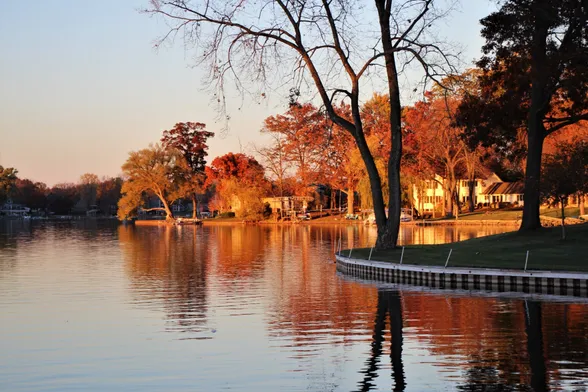 Autumn sunset on a lake with a few trees in dark silhouette in front of a background of trees in fall color lit by a setting sun.