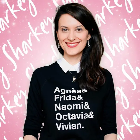 a profile photo of a woman with long dark hair wearing a black sweatshirt that says "Agnès & Frida & Naomi & Octavia & Vivian". The background is bubblegum pink and there is the word Sharkey appearing multiple times as a pattern