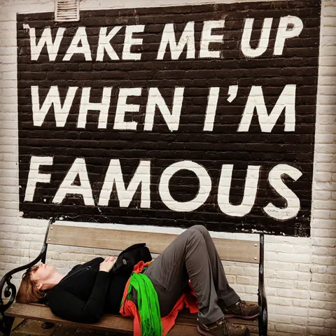 Alison is lying on a bench under a sign on a brick wall that says "Wake me up when I'm famous" in large white letters on a black background.