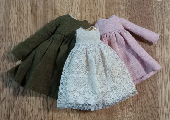 Three doll-sized dresses laid out on a wooden table. Two with long sleeves, one in green linen and one in pink linen. And another dress with no sleeves in light cream linen and lace.