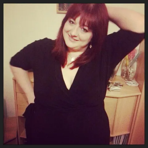 Mel, smiling, stands in front of a living room cabinet. She has long, red dyed hair with bangs, and wears a black dress. One arm is lifted behind her head, the other one pressed against her hip. 