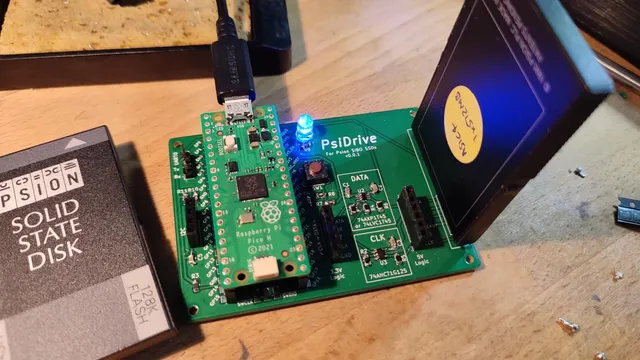 Photo of a fully constructed PsiDrive 0.0.1, including a Raspberry Pi Pico mounted on top. There is a SIBO solid state disk (SSD) plugged into the 6 SIBO-SP pins. A microUSB cable is plugged into the Pico. To the left of the PsiDrive is 128K Flash Psion SSD.